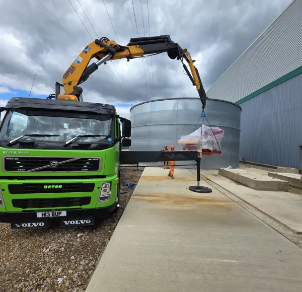 Green HIAB lorry lifting wrapped machinery beside a steel tank at an industrial site