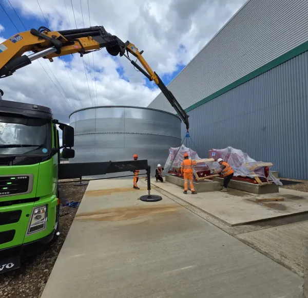 Green HIAB lorry lifting wrapped machinery beside steel tanks