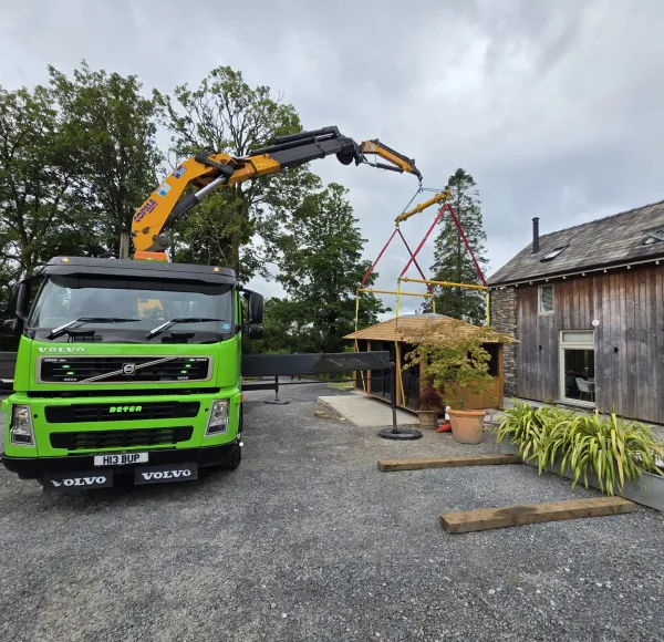 Green HIAB lorry lifting a garden building beside a barn